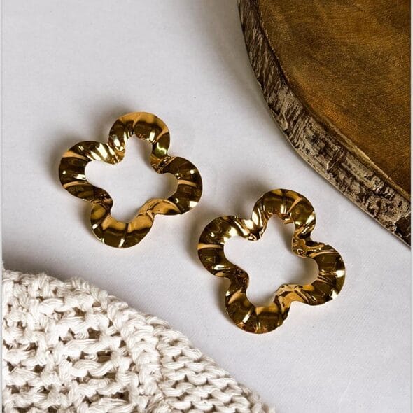 Pair of gold-plated floral statement earrings with wavy edges, displayed on a white surface next to a rustic wooden board and a knitted fabric.