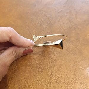 Gold-plated geometric bangle bracelet with two triangle-shaped ends, displayed on a light brown textured surface and held by a hand.
