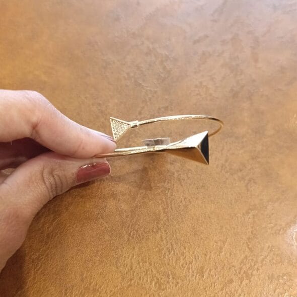 Gold-plated geometric bangle bracelet with two triangle-shaped ends, displayed on a light brown textured surface and held by a hand.