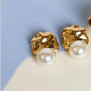 Pair of Gold and Pearl Statement Earrings with sculpted design and large faux pearl accents, displayed on a two-tone background of light beige and pastel blue.