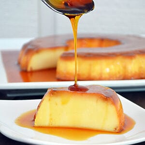 Brazilian milk flan (pudim) from Karamelado, displayed on a white plate with glossy caramel sauce dripping down the sides. The dessert has a smooth, golden surface and is surrounded by festive Christmas decorations such as pine branches and ornaments, creating a cozy holiday vibe