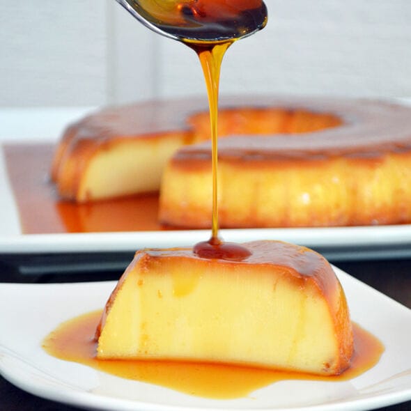 Brazilian milk flan (pudim) from Karamelado, displayed on a white plate with glossy caramel sauce dripping down the sides. The dessert has a smooth, golden surface and is surrounded by festive Christmas decorations such as pine branches and ornaments, creating a cozy holiday vibe