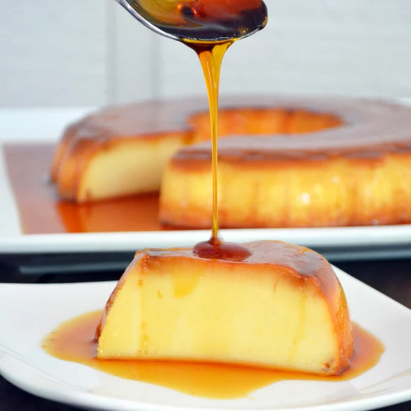 Brazilian milk flan (pudim) from Karamelado, displayed on a white plate with glossy caramel sauce dripping down the sides. The dessert has a smooth, golden surface and is surrounded by festive Christmas decorations such as pine branches and ornaments, creating a cozy holiday vibe