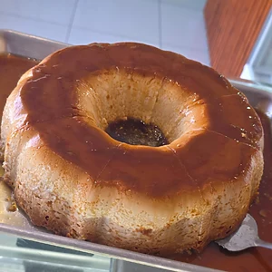 Large Brazilian milk flan (pudim) from Karamelado displayed on a white plate, topped with glossy caramel sauce dripping down the sides. The dessert has a smooth golden surface and is surrounded by festive Christmas decorations such as pine branches, ornaments, and warm lights, making it a perfect holiday centerpiece.