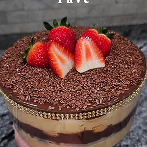Holiday-themed image featuring Karamelado’s Double Brigadeiro Pavê in a clear glass bowl. The dessert shows layers of white brigadeiro cream, chocolate brigadeiro, and crushed biscuits, topped with chocolate sprinkles and three fresh strawberries. Festive pine branches and ornaments decorate the background, with text reading: ‘The showstopper is back – That dreamy pavê in a gorgeous clear bowl.