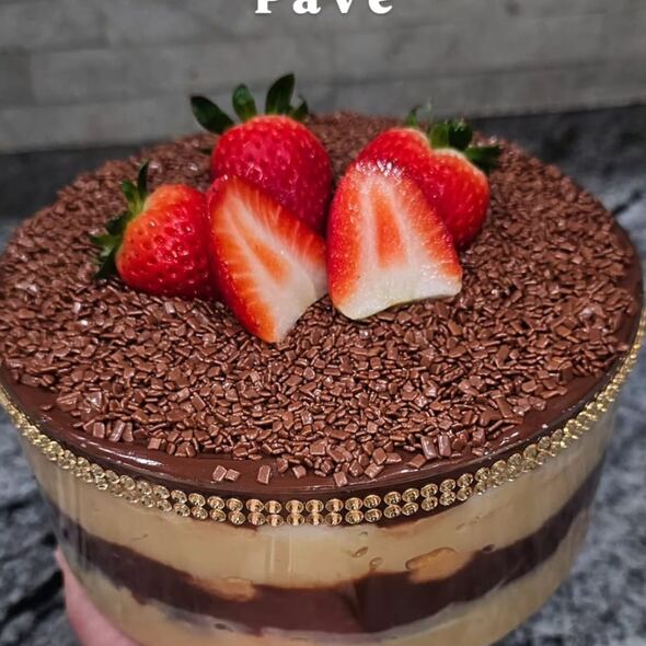 Holiday-themed image featuring Karamelado’s Double Brigadeiro Pavê in a clear glass bowl. The dessert shows layers of white brigadeiro cream, chocolate brigadeiro, and crushed biscuits, topped with chocolate sprinkles and three fresh strawberries. Festive pine branches and ornaments decorate the background, with text reading: ‘The showstopper is back – That dreamy pavê in a gorgeous clear bowl.