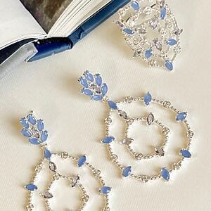 Elegant silver-tone jewelry set with circular openwork designs and marquise-cut blue sapphire stones, displayed on a book background, representing timeless elegance and rare beauty by Madu Joia Rara.