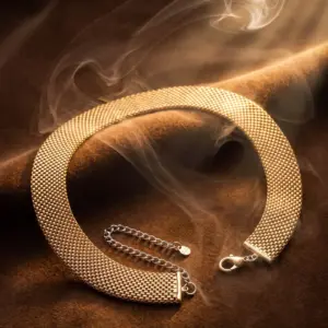 Wide gold-tone mesh collar necklace with a flexible woven design and adjustable chain extension, displayed on a luxurious warm background, representing modern elegance and timeless sophistication by Madu Joia Rara.