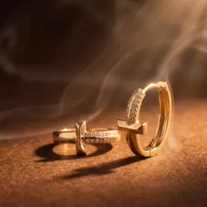 Golden Crossroads Zirconia Hoops Gold-tone hoop earrings featuring a modern crossbar design and sparkling zirconia stones, displayed on a warm golden background with soft swirling light, showcasing contemporary elegance by Madu Joia Rara.