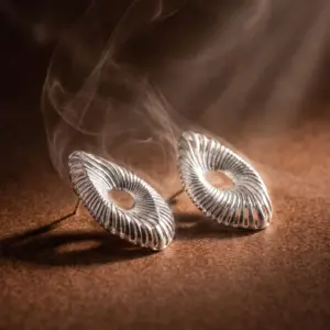 Silver-tone statement earrings featuring an open-center oval design with textured radiating lines inspired by ocean waves, showcasing modern elegance and artistic craftsmanship by Madu Joia Rara.