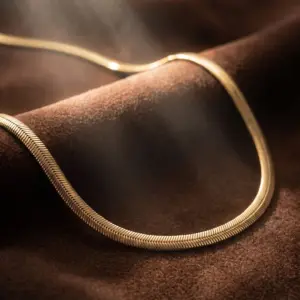 Elegant gold-tone herringbone necklace with a smooth flat design and luminous finish, displayed on a soft brown velvet surface, showcasing timeless sophistication by Madu Joia Rara.